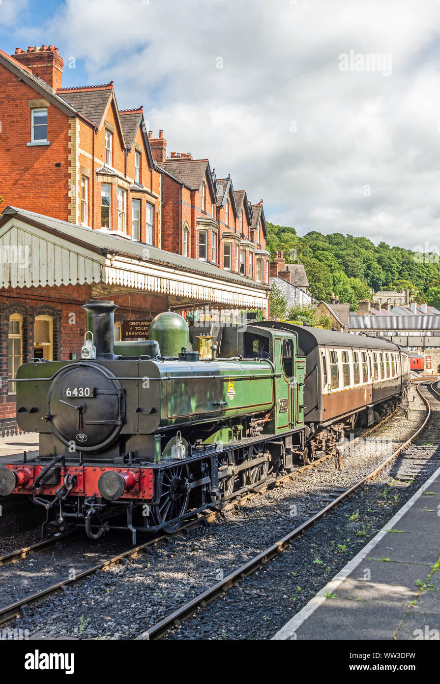 In tema di Llangollen Railway Station. Una vecchia locomotiva a vapore e carrozze a riposare accanto a una piattaforma pronta per uscire. Un bosco è in distanza. Foto Stock