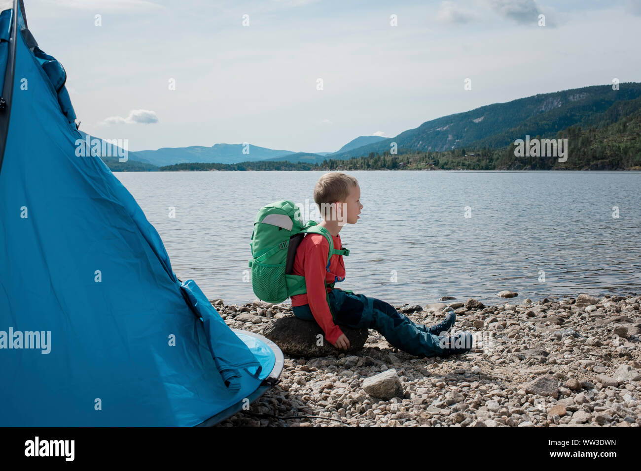 Ragazzo giovane camping seduto su una roccia con uno zaino e una tenda Foto Stock