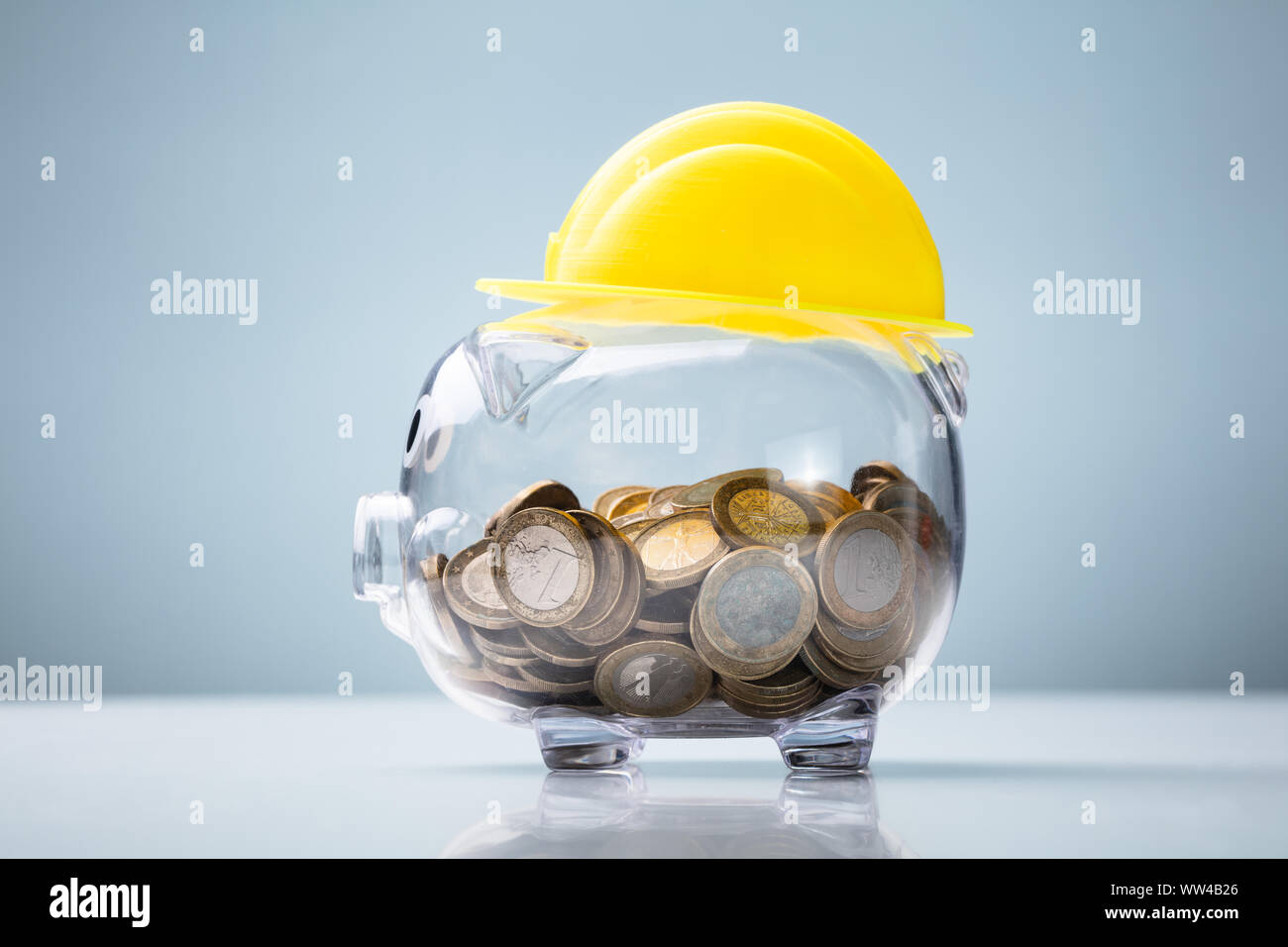 Close-up di Casco giallo su trasparente di salvadanaio sulla scrivania bianca contro lo sfondo blu Foto Stock