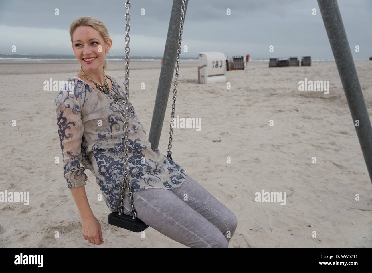 Ritratto di metà donna sulla spiaggia seduta su swing con sdraio in spiaggia e mare in background Foto Stock