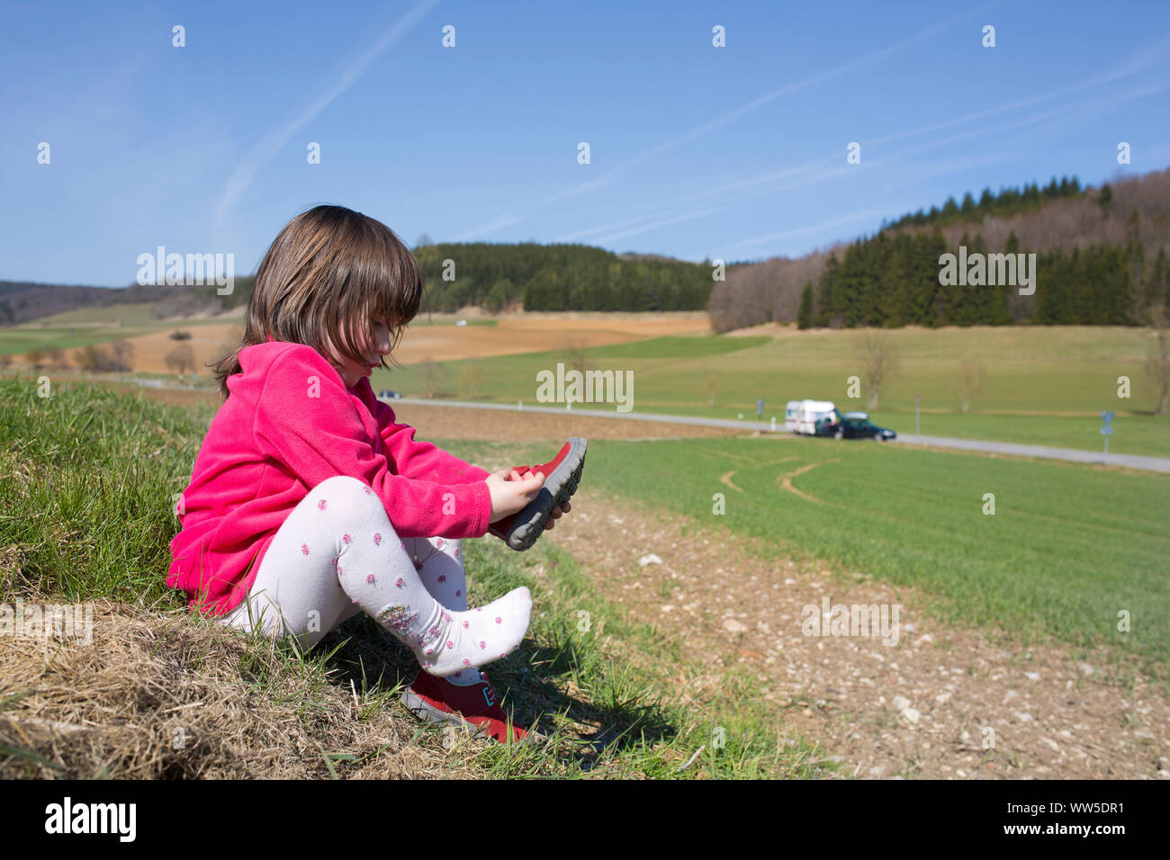 5-anno-vecchia ragazza con giacca rossa seduta al fieldedge, mettendo sulle scarpe Foto Stock