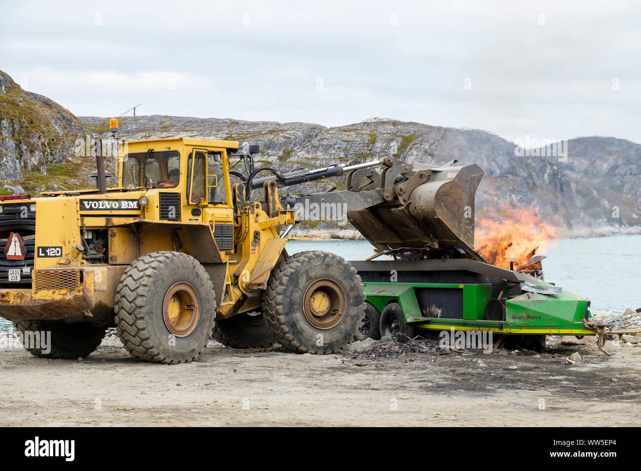 Un escavatore immondizia di dumping in un BurnBoss bruciando i rifiuti presso il locale punta. Paamiut (Frederikshåb), Sermersooq, Groenlandia Foto Stock