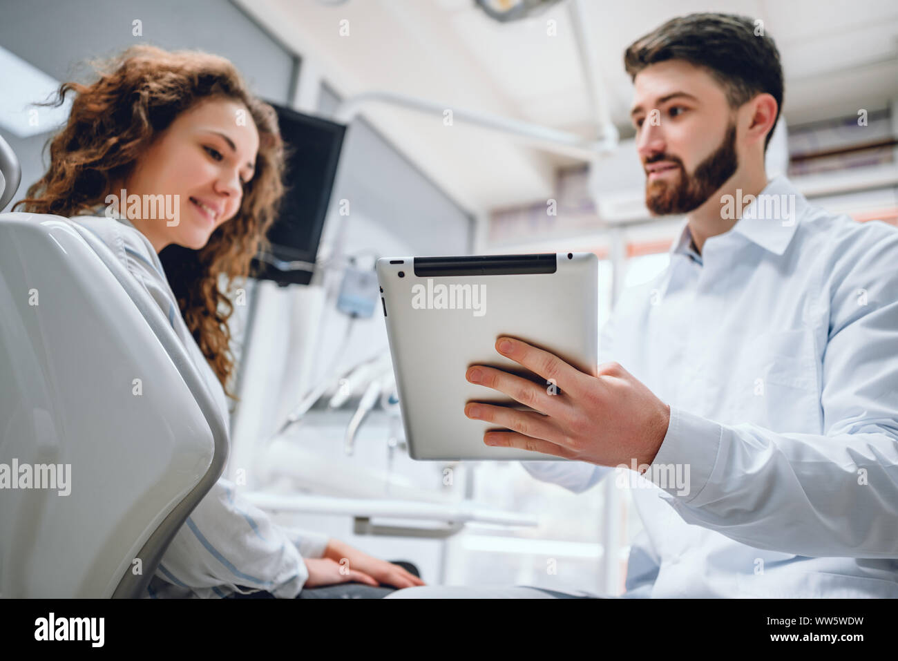 Felice dentista e paziente commentando trattamenti in un tablet applicazione in una consultazione con attrezzature mediche in background. Foto Stock