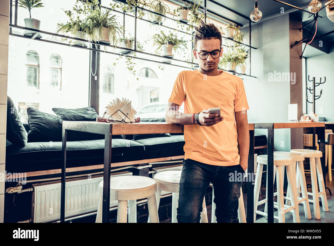 Giovane maschio freelance indiano in un fast food cafe, uomo bello la lettura di un messaggio di testo durante il lavoro sul telefono cellulare in confortevole coffee shop. Vista panoramica wi Foto Stock