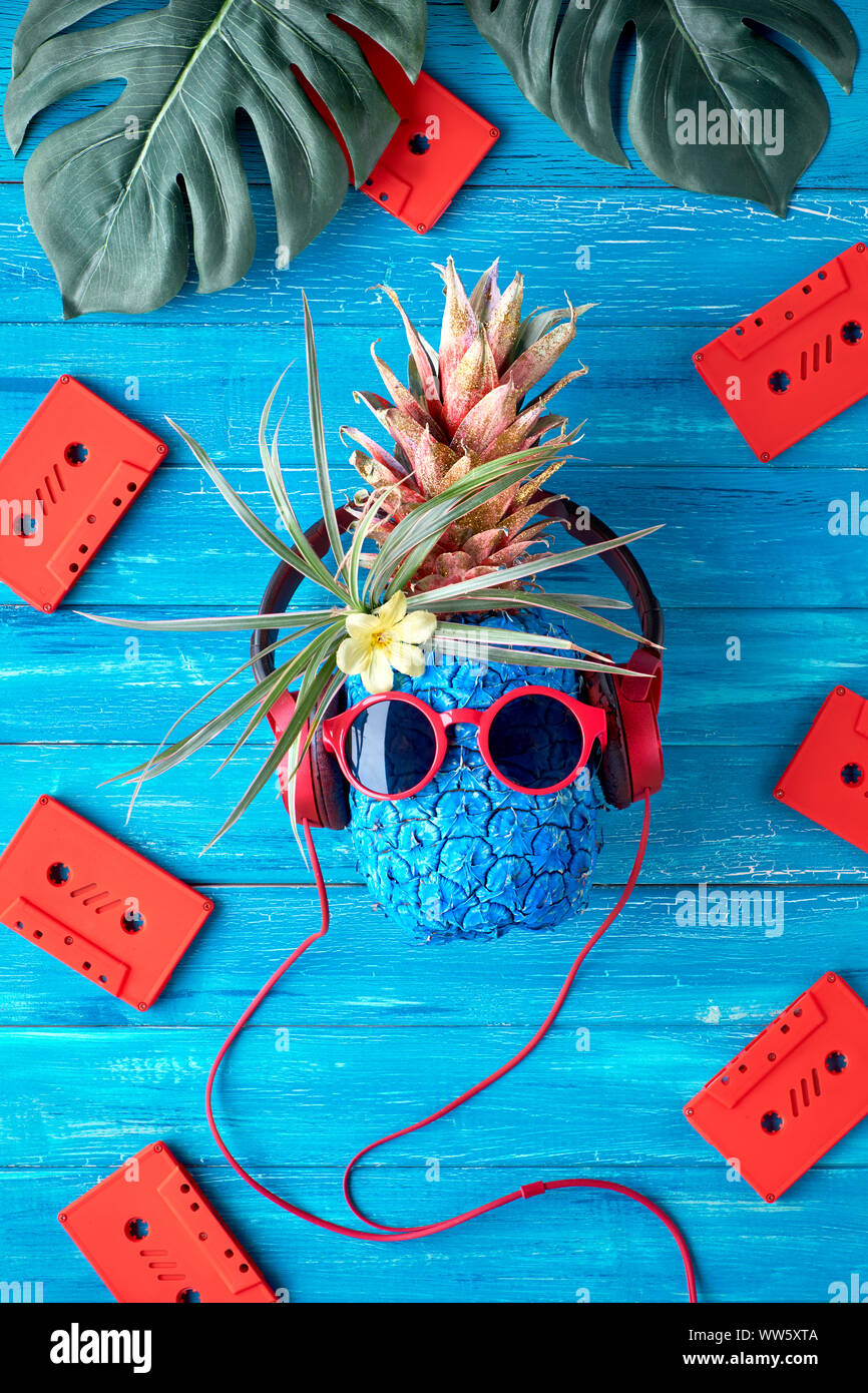 Tanga carattere di ananas in rosso gli occhiali da sole e auricolari ascolta la musica. Rustico in legno a trama sfondo blu con red cassette audio e Foto Stock