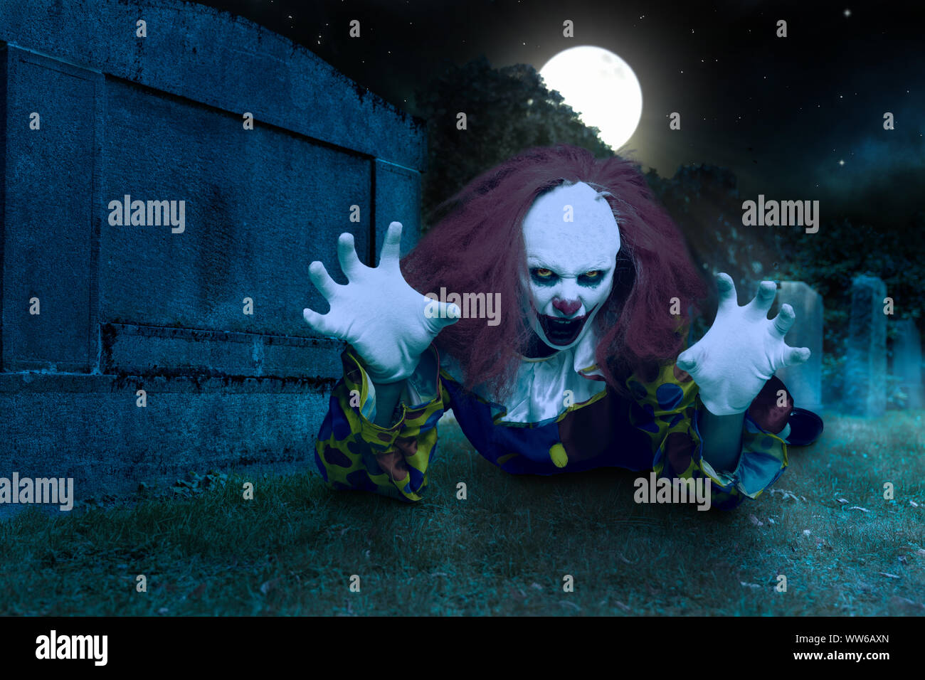 Un pauroso clown si trova di fronte a una lapide funeraria e fa un gesto spaventoso Foto Stock