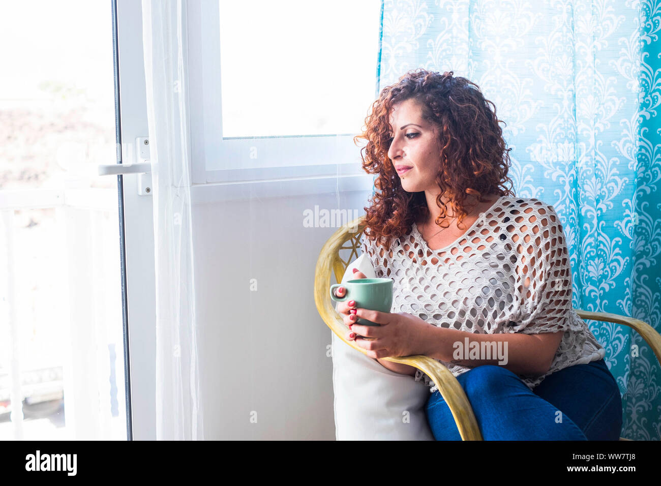 Una donna in casa vicino alla finestra con una tazza di tè o caffè. luce brillante e pensieri per la bellissima femmina in una casa chiara. Foto Stock