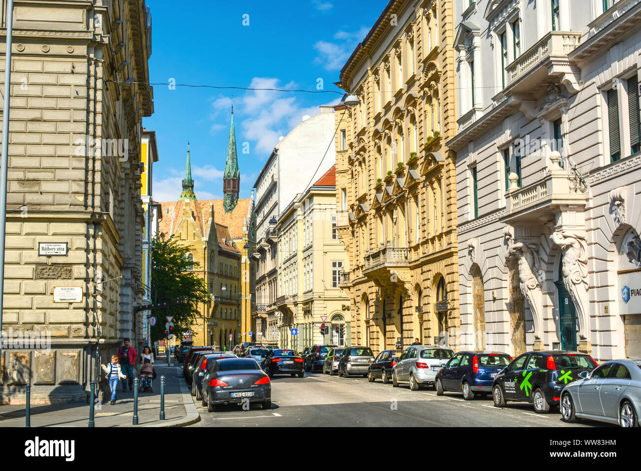 Una famiglia passeggiate passato auto fuori di Bathory Utca, o Bathory street nel distretto V di Budapest in Ungheria, con la Chiesa Unitaria guglie della distanza Foto Stock