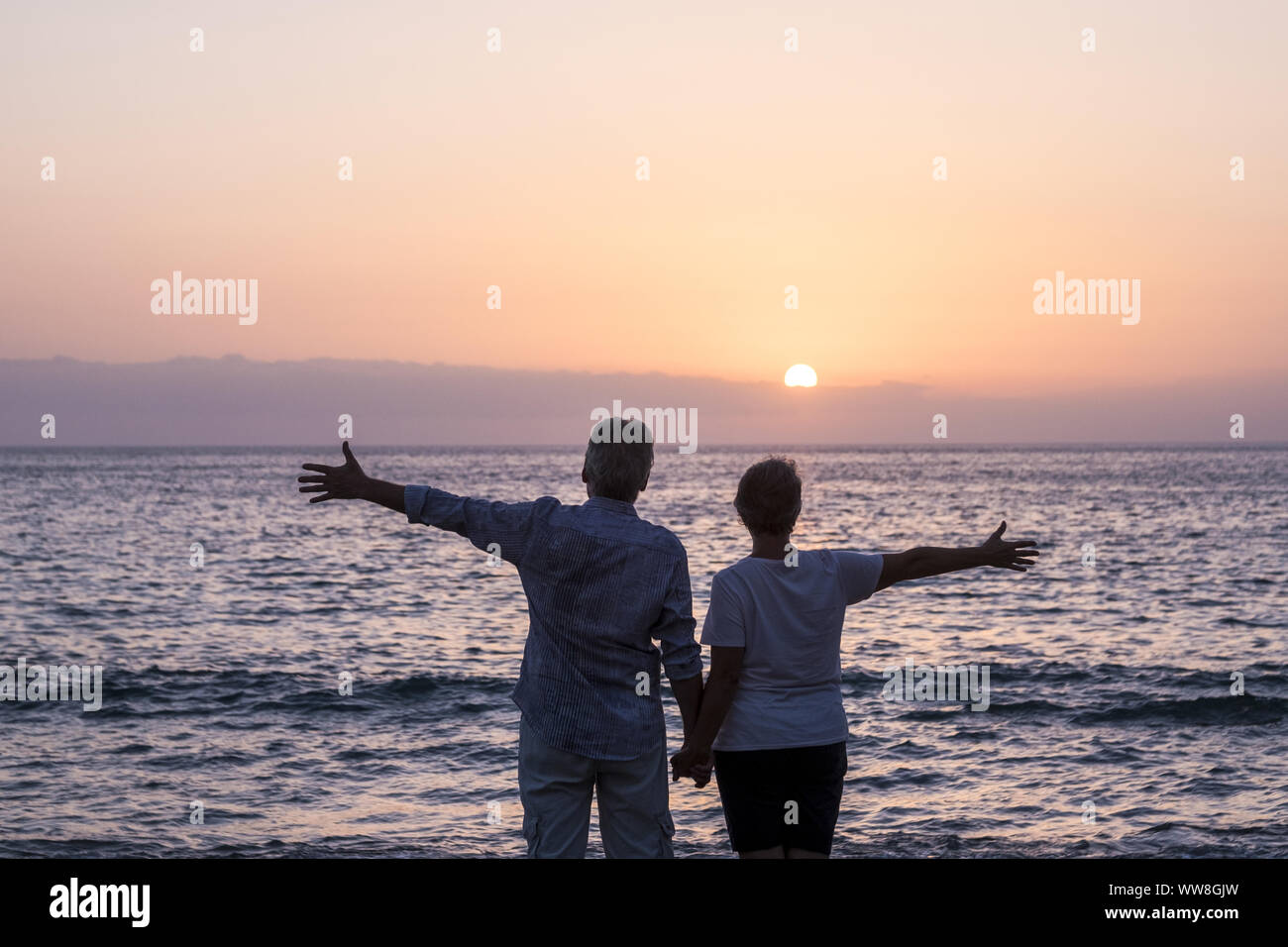 Concetto di vacanza, turismo, viaggi e persone - felice coppia senior sulla spiaggia ghiaiosa di ridere e scherzare guardando il mare al tramonto con le braccia aperte, i capelli bianchi Foto Stock