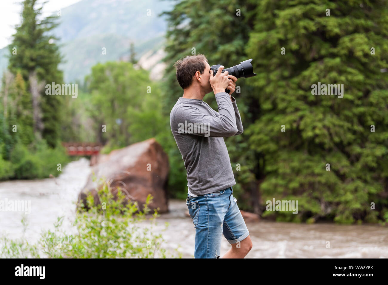 Uomo di scattare le foto con la fotocamera in Redstone, Colorado durante la stagione estiva con un grande masso e ponte rosso sul fiume di cristallo da alberi Foto Stock