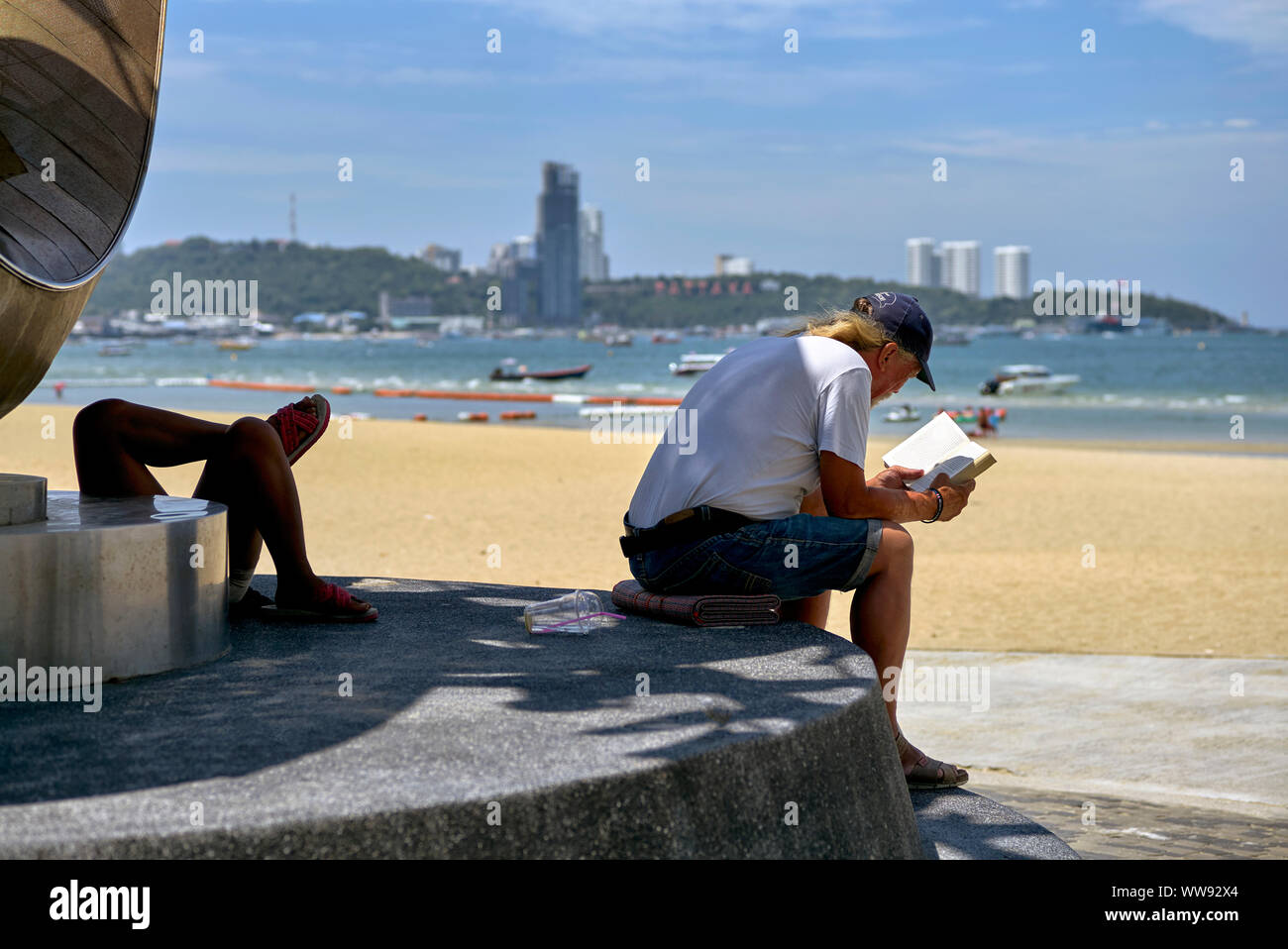 Persone che si rilassano all'aperto in varie forme in spiaggia con un uomo che legge un libro e compagno sdraiato a croce zampe. Thailandia Sud-est asiatico Foto Stock