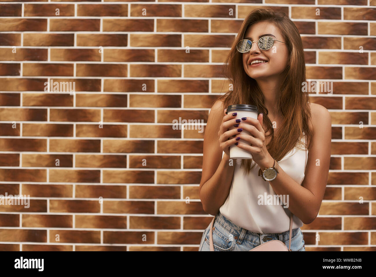 Ritratto di Allegro giovane donna in occhiali da sole in piedi su sfondo mattone durante il photoshoot per esterni. Lei è alla ricerca di distanza con un sorriso mentre si tiene il suo drink da asporto. Inquadratura orizzontale Foto Stock