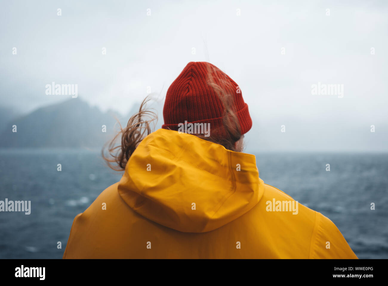 Hipster traveler indossando Red Hat e impermeabile giallo che guarda lontano a nuvoloso montagna e mare tempestoso. Da solo uomo che viaggia a scandinavian autentica la Foto Stock
