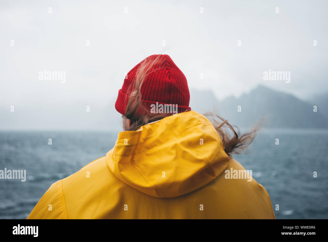 Viaggiatore Da Solo indossando un impermeabile giallo e Red Hat guardando il mare tempestoso e nebbioso montagna. Stile di vita outdoor adventure, scandinavo wanderlust Foto Stock
