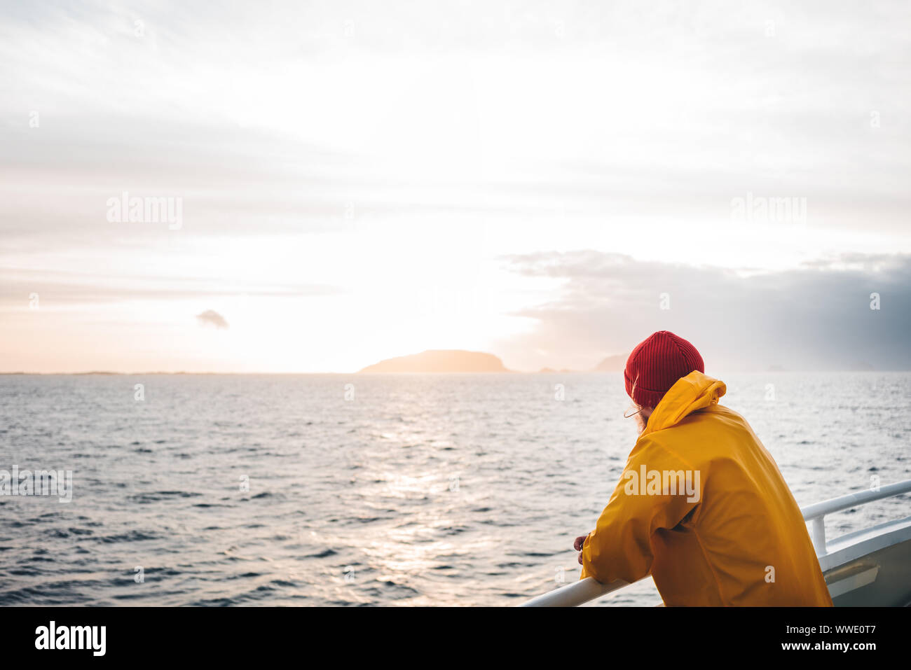 Giovani traveler indossando cappello rosso e giallo impermeabile galleggiante sulla nave guardando il tramonto mare dopo la tempesta e le montagne di nebbia sulla skyline. Lifestyle travel Foto Stock