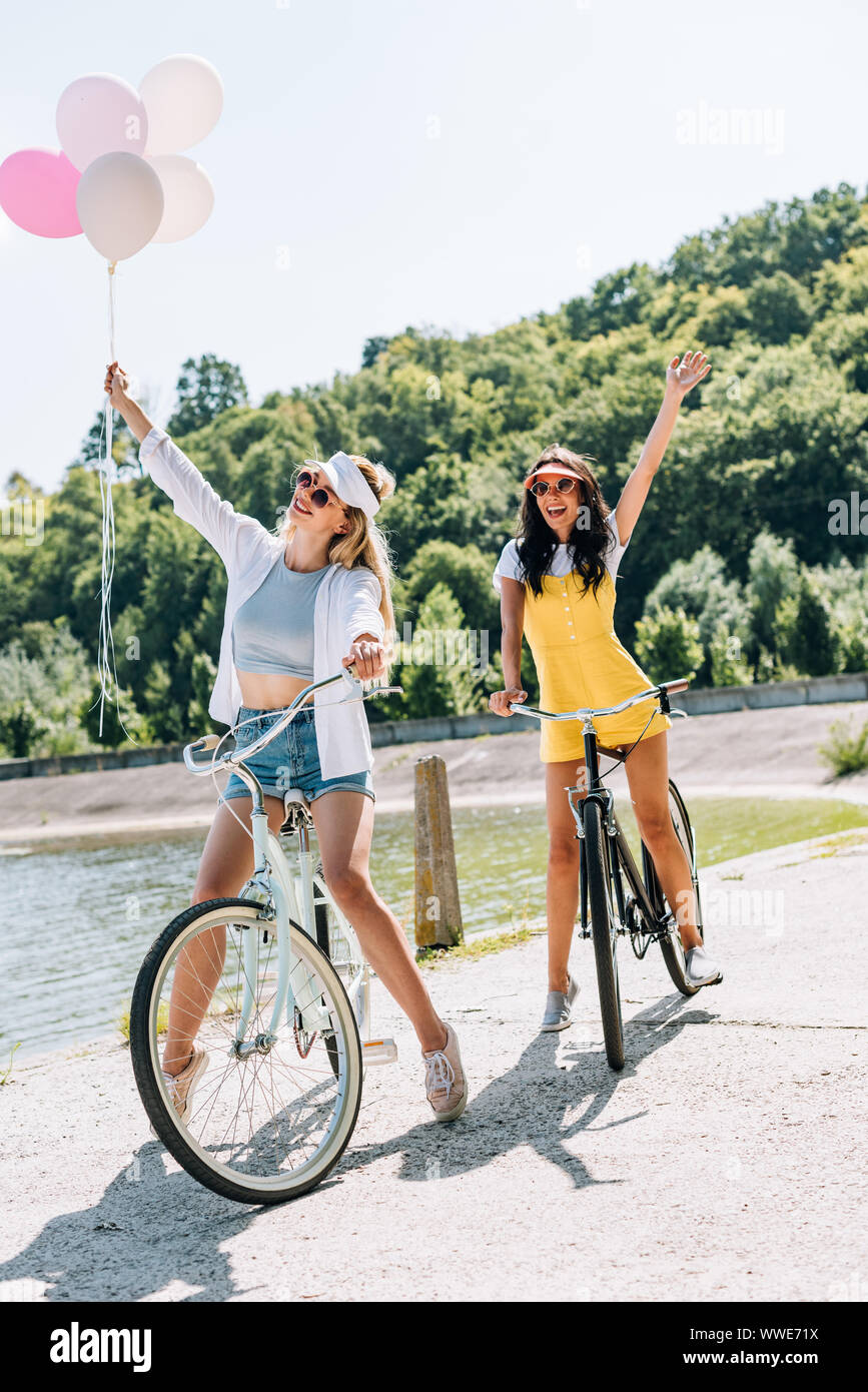 Felice bionda e bruna amici equitazione biciclette con palloncini vicino al fiume in estate Foto Stock