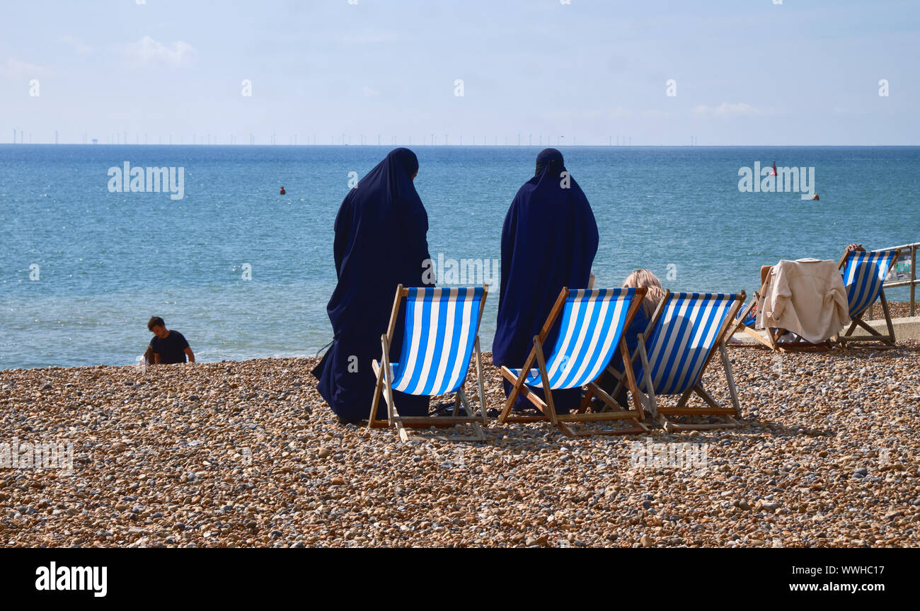 Due donne musulmane permanente sulla rocciosa spiaggia di Brighton circa di sedersi sulla sedia a sdraio accanto a loro capelli biondi amico come si vede dalla parte superiore Foto Stock