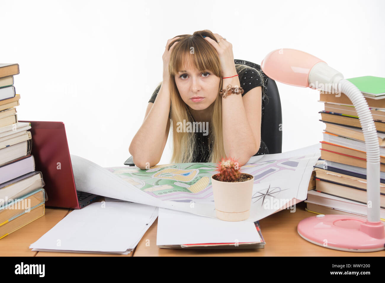 Studente designer dietro una scrivania con una pila di libri e di progetti si siede tenendo la testa tra le mani Foto Stock