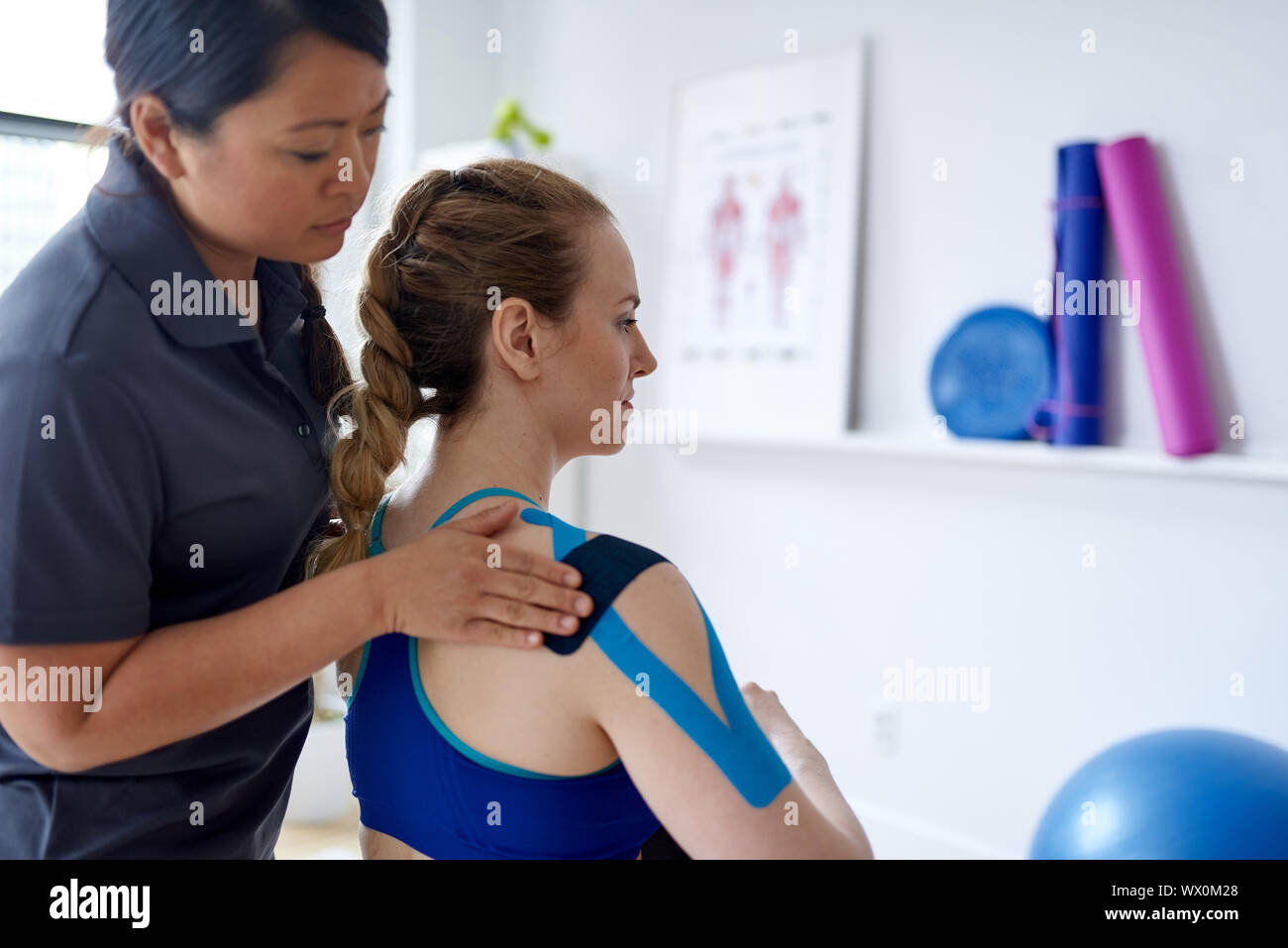 Donna cinese massaggi terapista applicazione kinesio tape per le spalle e il collo di un attraente bionda client in un luminoso ufficio medico Foto Stock