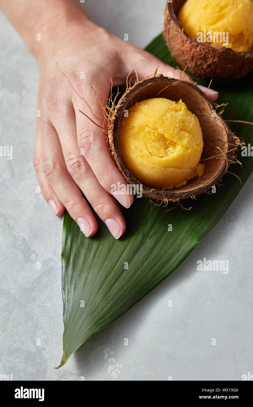 Un gelato al mango sorbetto in un guscio di noce di cocco è tenuto da una femmina di mano su una foglia di palma su un cemento grigio Sfondo. Vista superiore Foto Stock