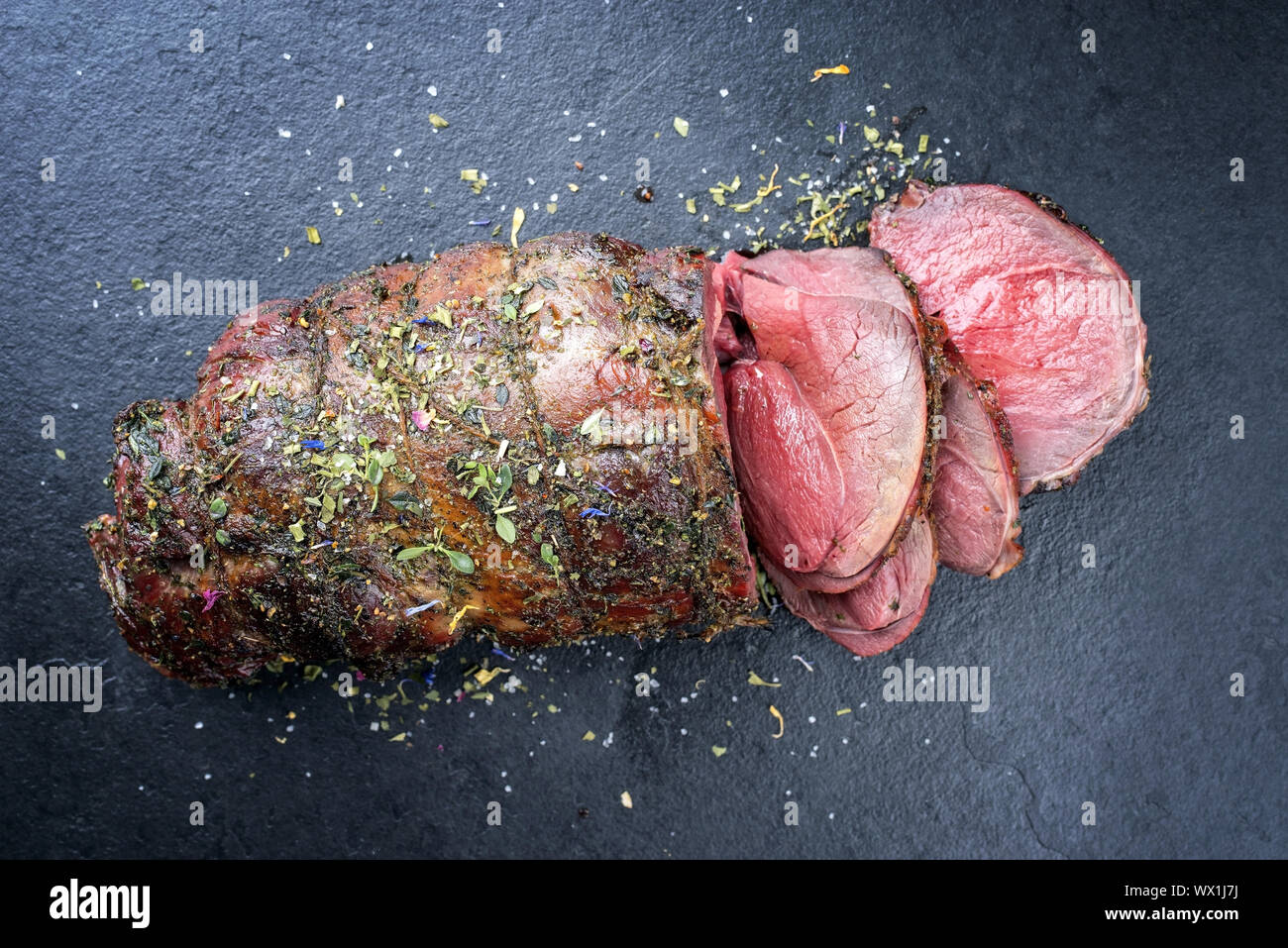 Barbecue secca haunch invecchiato di cervo con erbe aromatiche come close-up su una scheda di colore nero Foto Stock