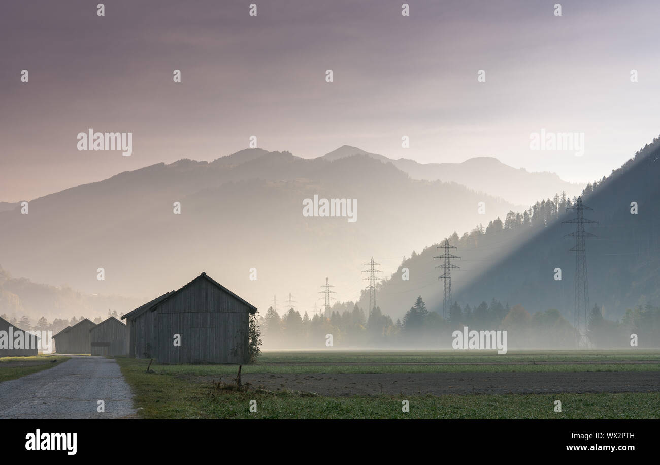 Nebbia di mattina in una valle di montagna con i campi e vecchi fienili in legno e croce di reticolo di linee di potenza e Foto Stock