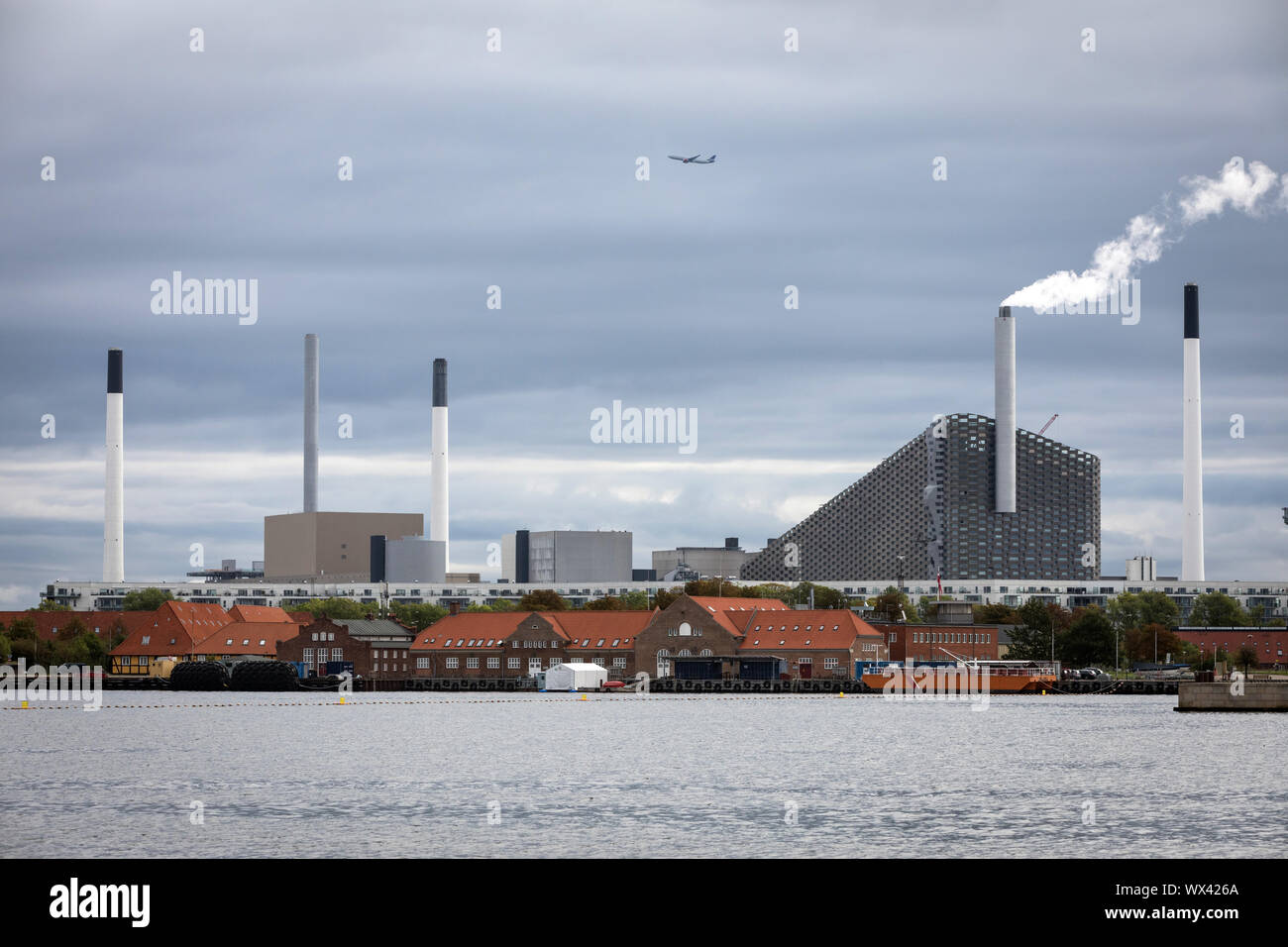 Un aereo sorvola l'Amager Bakke combinati inceneritore e pista da sci a Copenhagen, in Danimarca, con il porto in primo piano Foto Stock