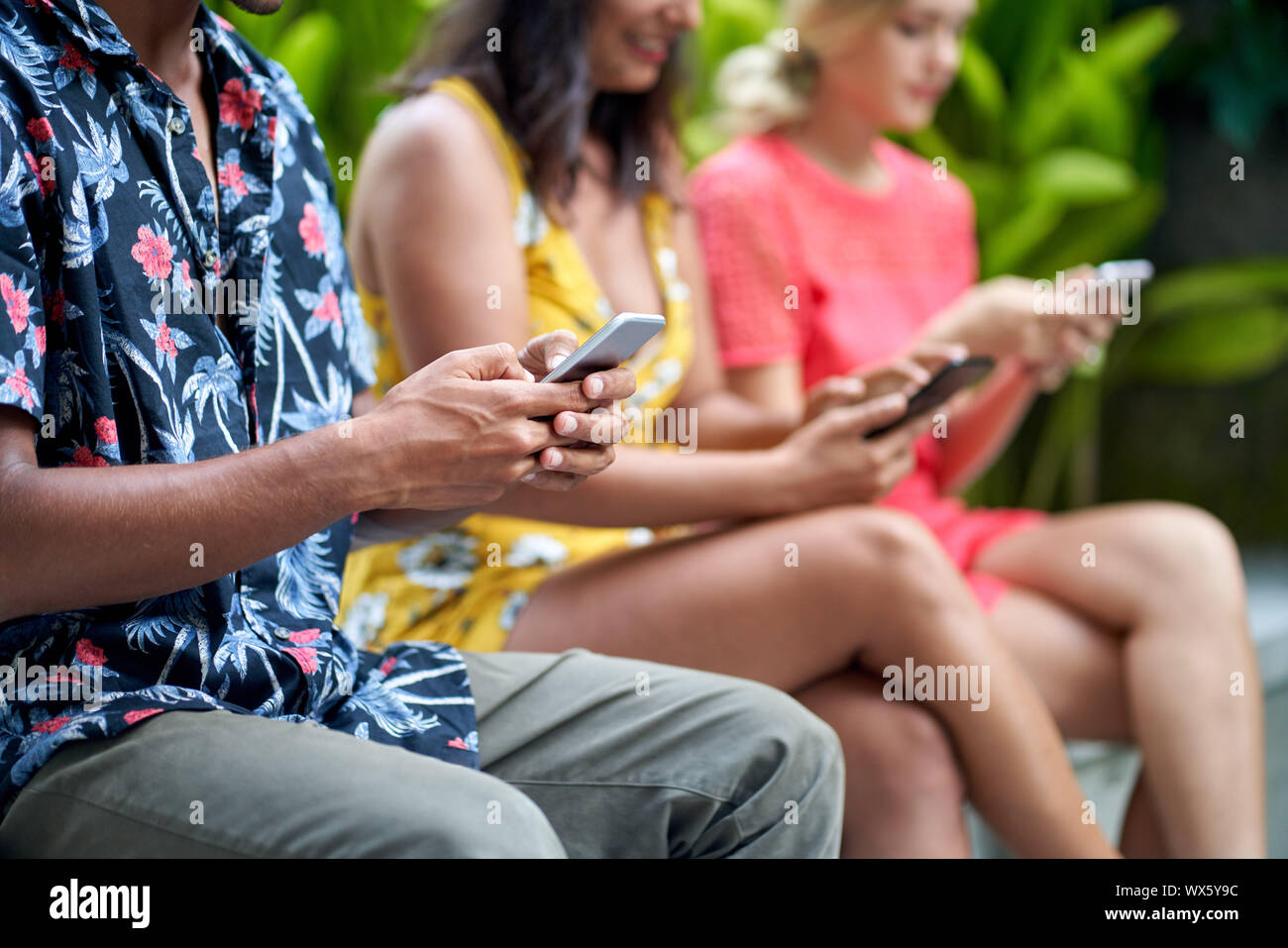 Unposed closeup lifestyle colpo di tre multi-etnico popolo millenario digitando su smartphone ina giardino tropicale con piante verdi Foto Stock