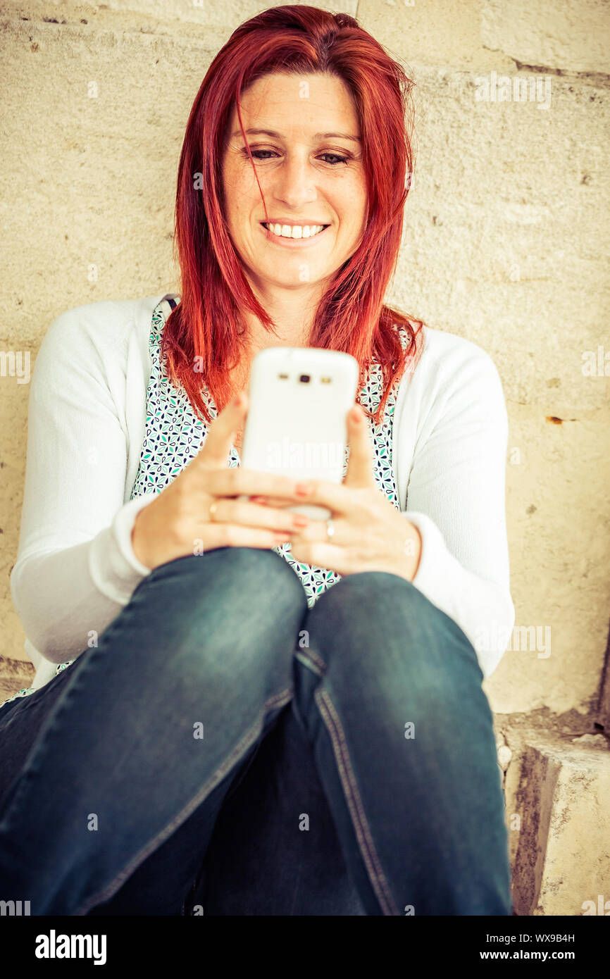 Piuttosto sorridente redhead donna seduta contro un vecchio muro nel processo di invio di messaggi mediante il telefono cellulare Foto Stock