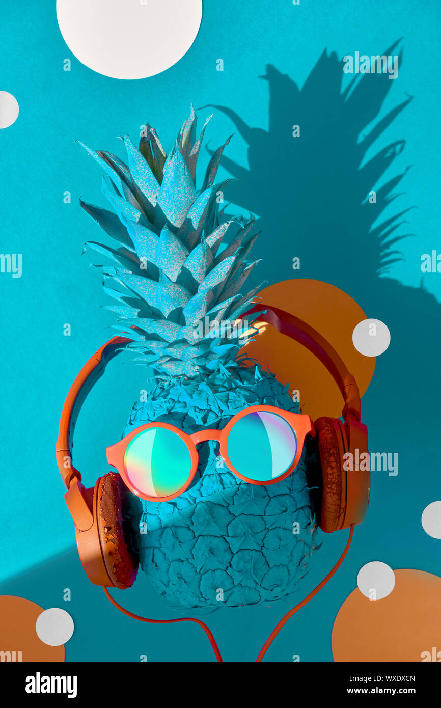 Funny ananas in occhiali da sole e auricolari con un'ombra, piatto adagiare sulla carta blu sullo sfondo Foto Stock
