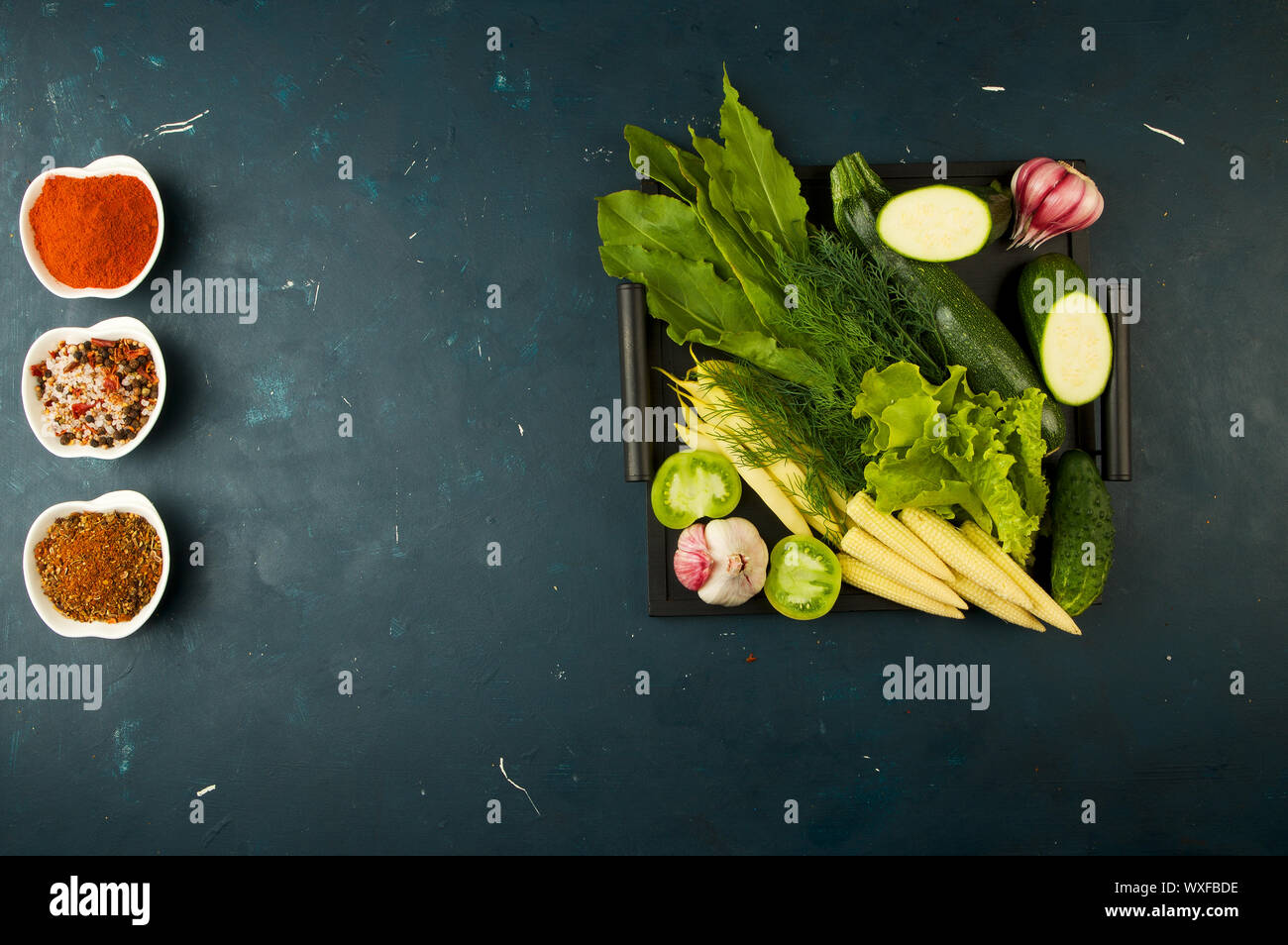 Le verdure in scatola sulla pietra uno sfondo scuro. Giovani Verdi aglio cipolla zucchine spezie luminose laici in un vassoio di legno Foto Stock