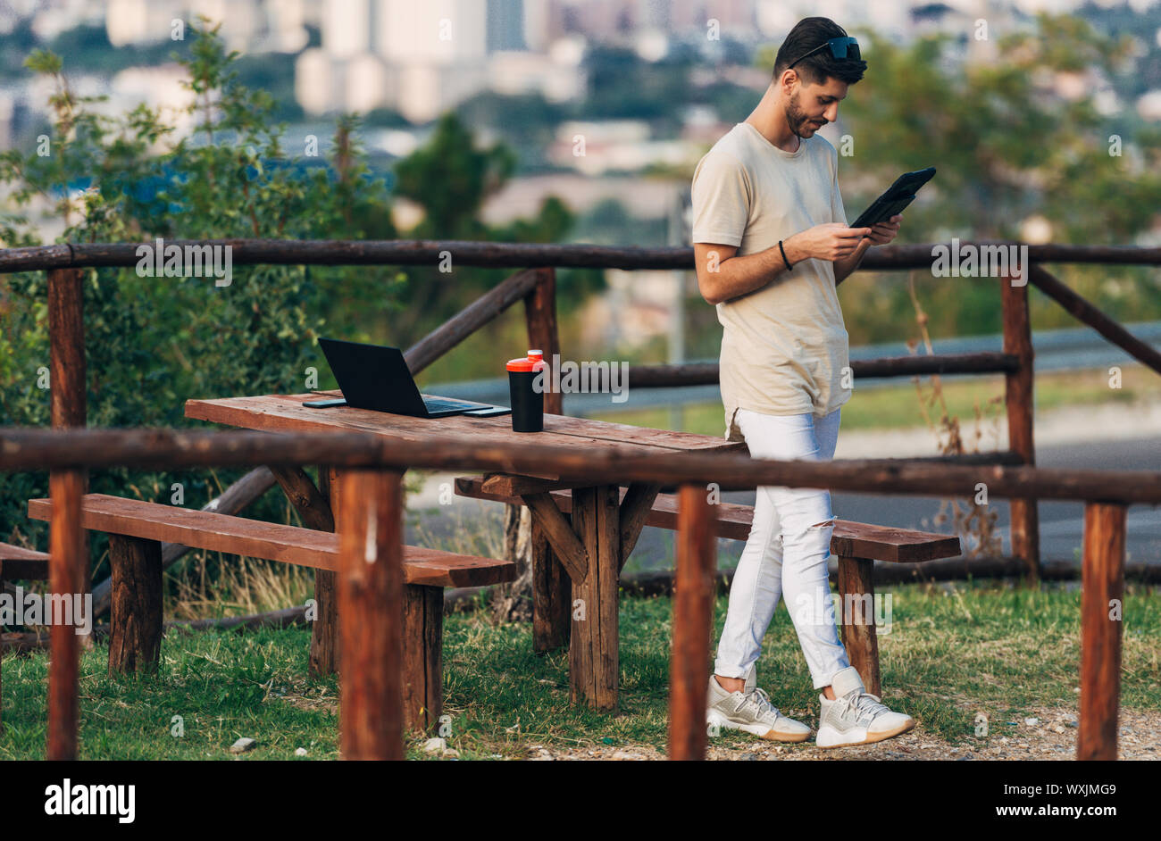 Bello il freelance in abito casual lavora con laptop e tablet in collina contro vista città Foto Stock