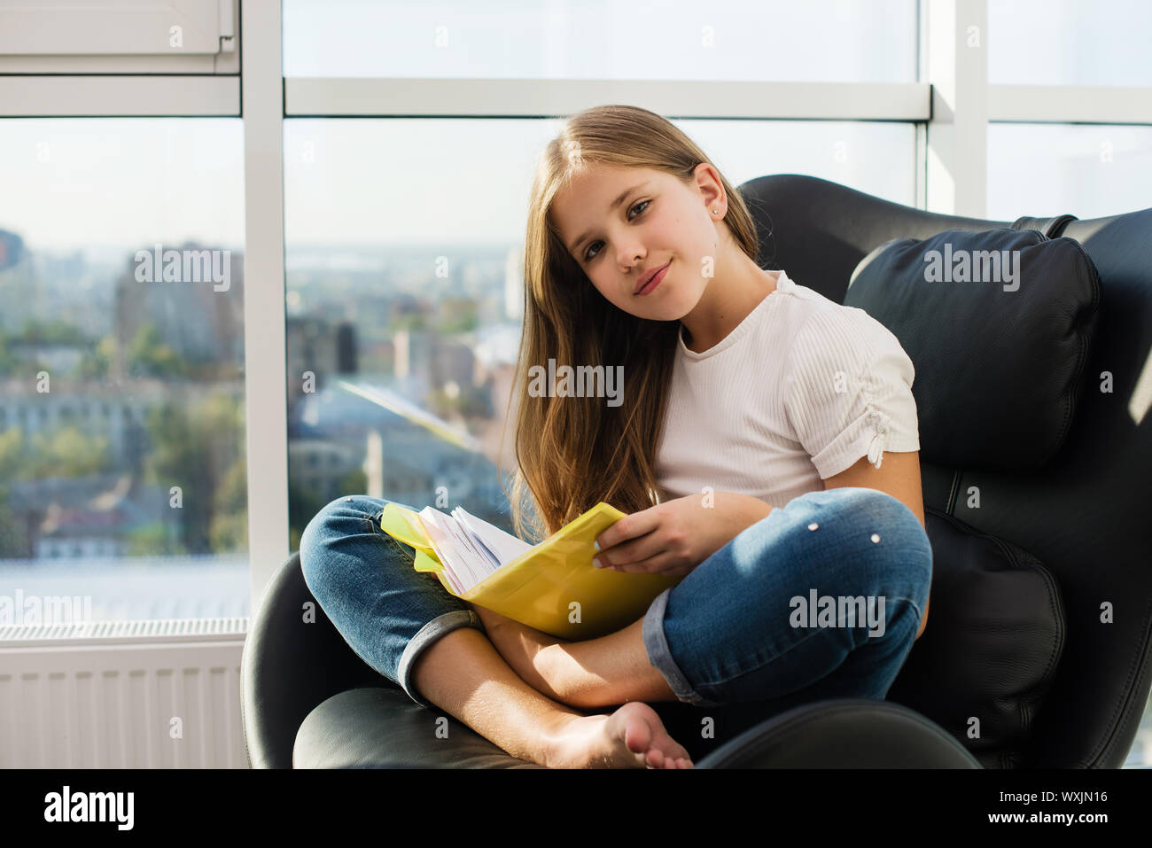 Preteen positivo studentessa in posa durante la lettura del libro di testo in copertina di colore giallo, seduta in lotus pongono in pelle nera sedia con finestre panoramiche che ho Foto Stock