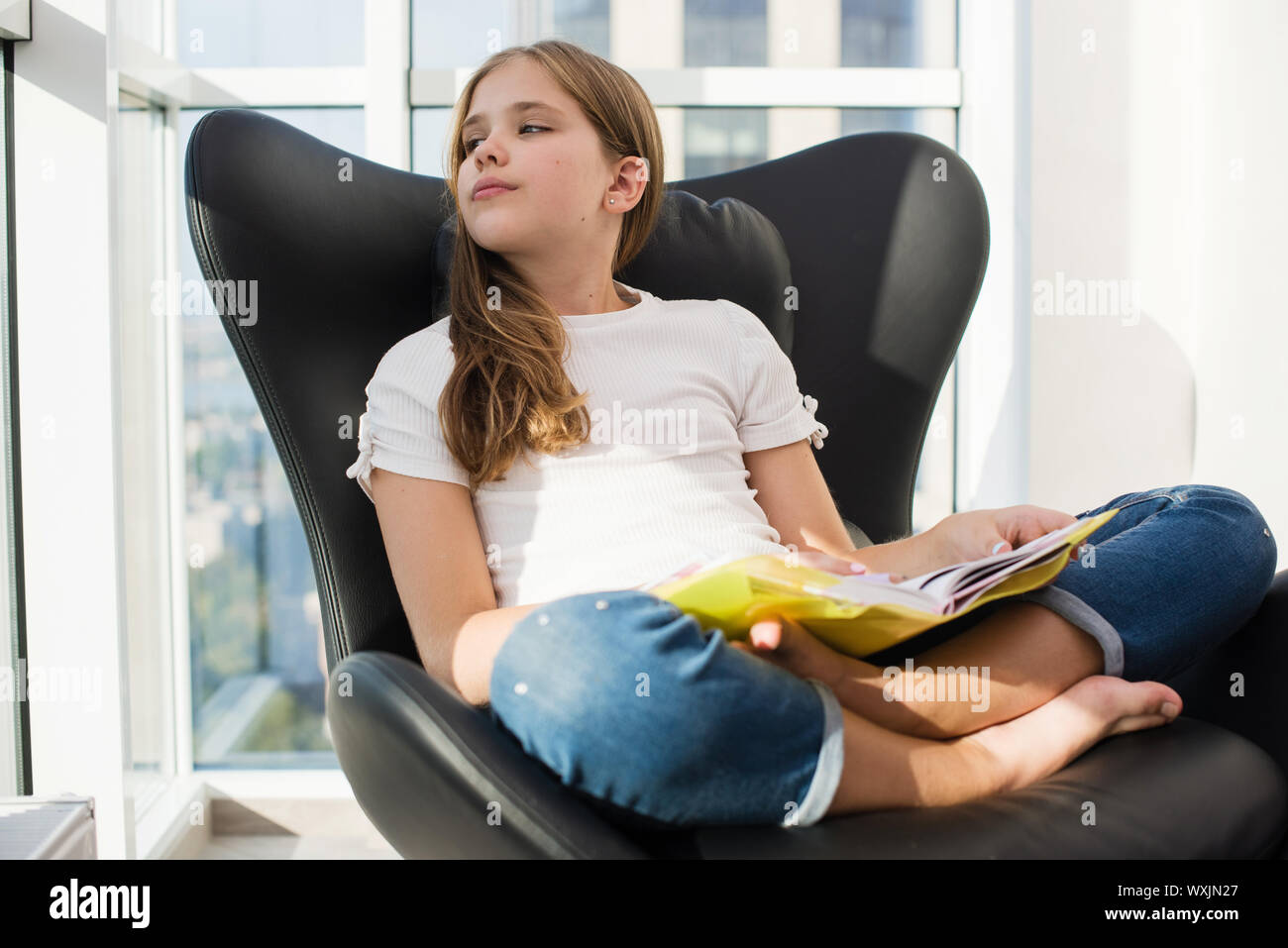 Dreaming preteen studentessa con la lettura del libro di testo nel coperchio giallo, seduta in lotus pongono in pelle nera sedia con finestre panoramiche interno, Foto Stock