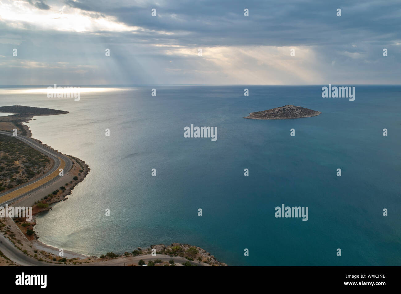 Inclinazione raggi solari rompere attraverso le nuvole alla superficie del mare. Isola nel mare e tortuosa strada di mattina. Vista aerea Foto Stock
