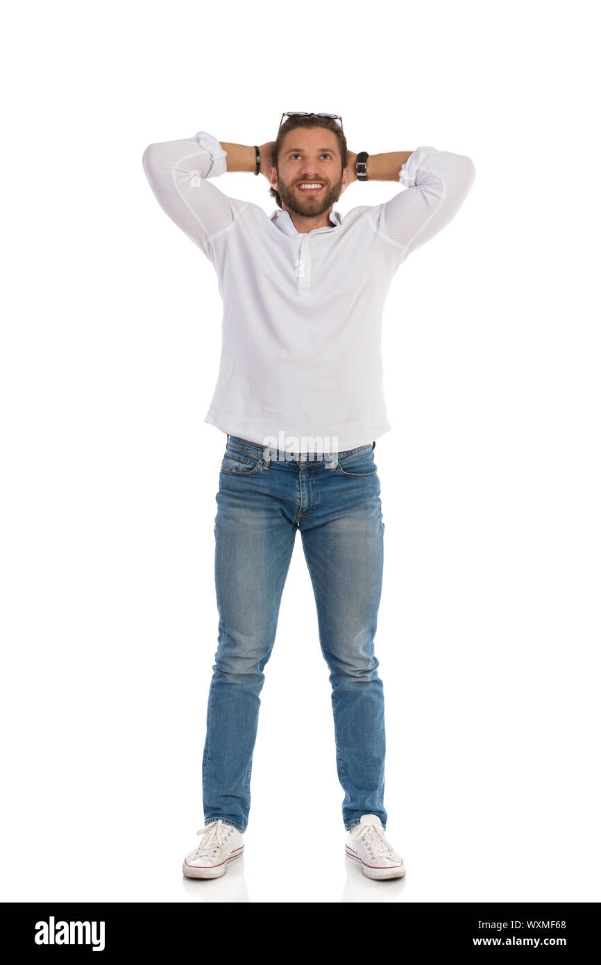 Bel uomo in jeans e scarpe da ginnastica e camicia bianca è rilassato permanente, tenendo le mani dietro la testa, sorridendo e guardando verso l'alto. Lunghezza completa studio shot isolat Foto Stock