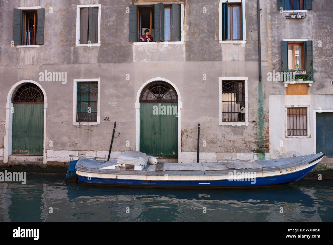 Canale di Venezia con una barca a motore d'acqua davanti a vecchi edifici e di una donna a finestra Foto Stock