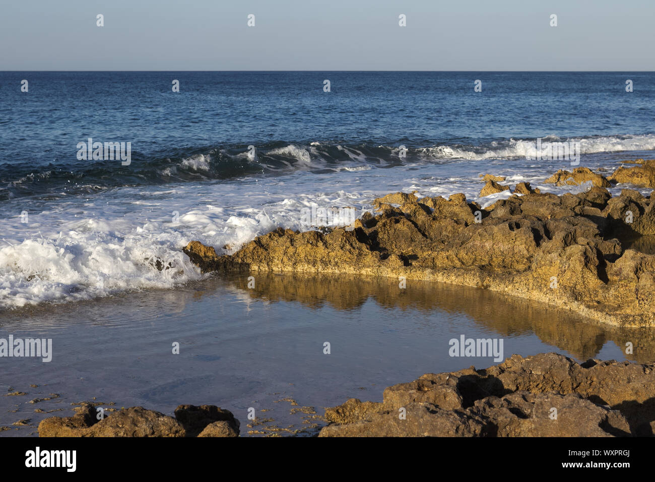 Acqua schiumosa di blu oceano Schiantati sulla spiaggia rocciosa illuminata da ora d'oro. Foto Stock