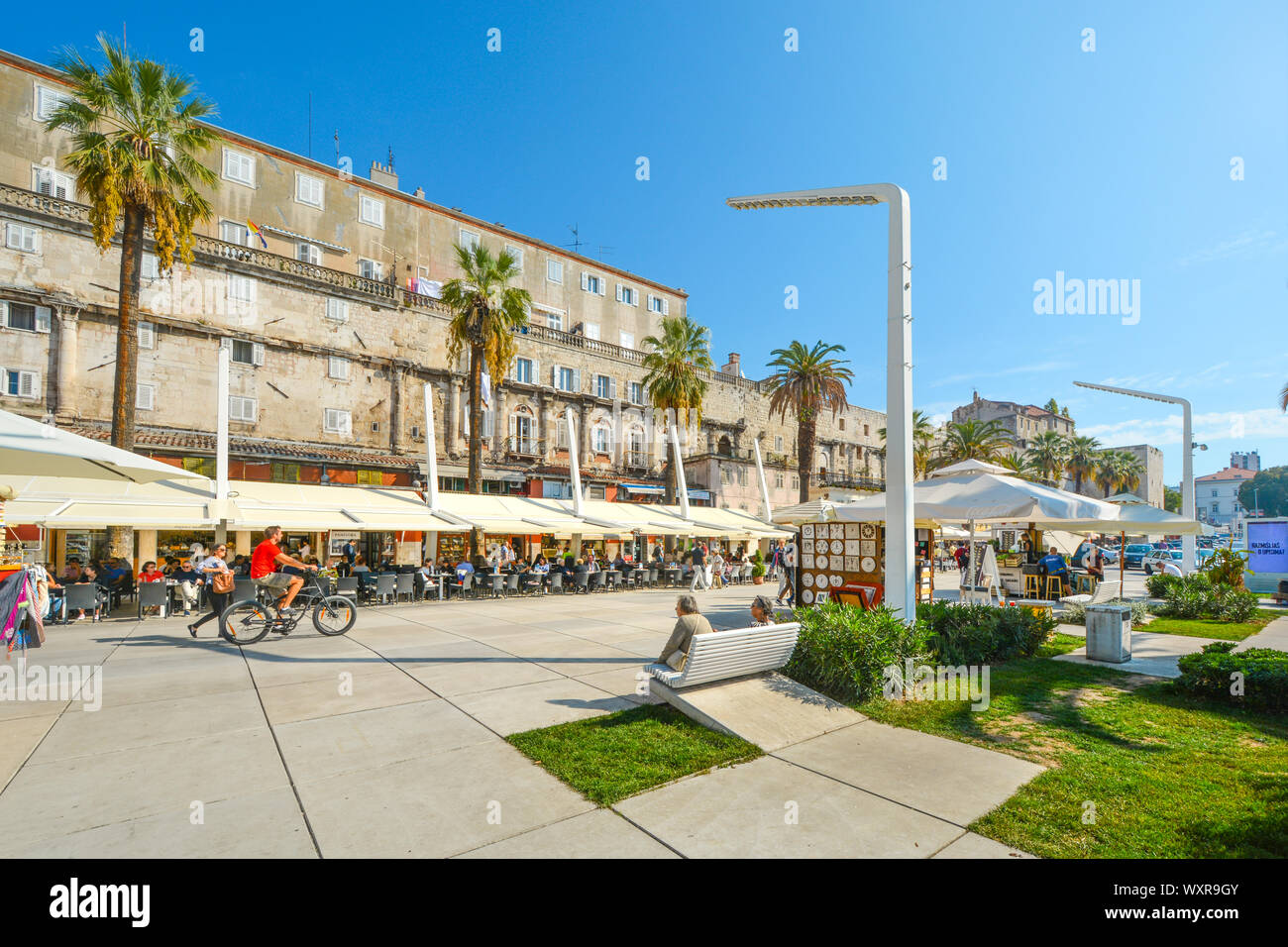 Pomeriggio estivo sul lungomare di Riva presso il porto di Spalato Croazia come turisti e un ciclista godere gli alberi di palme e caffetterie sulla costa Foto Stock