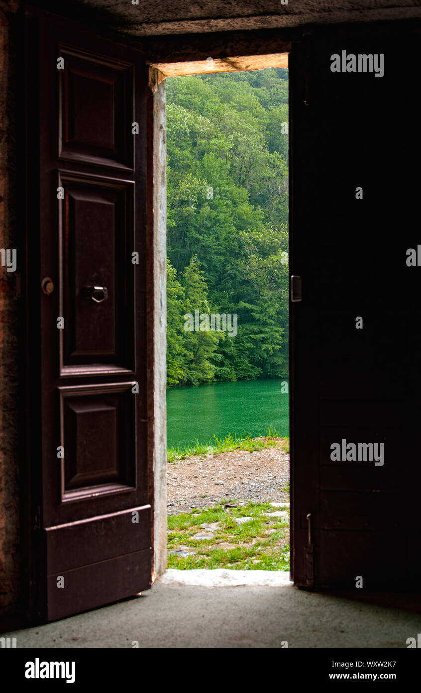 Aprire a doppia porta di legno di un edificio rurale affacciato su un tranquillo fiume o lago con alberi verdi intravisto attraverso l'apertura Foto Stock
