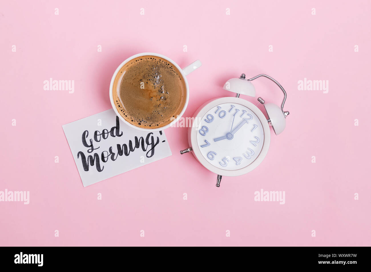 Analog sveglia, una tazza di caffè e nota con testo Buongiorno su sfondo rosa. Inizio del giorno concetto. Foto Stock