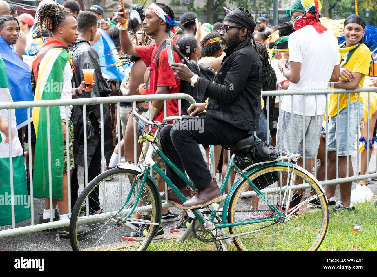 Hinter Zaun dem, un welcher die West Indian Day Parade entlang geht, sitzt ein Mann auf seinem Fahhrad und macht Fotos mit seinem Handy von den bunt un Foto Stock