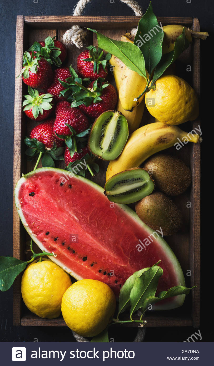 Varie colorata frutta tropicale selezione nel vassoio di legno su sfondo scuro, vista dall'alto. Anguria, fragola, limoni e kiwi Foto Stock